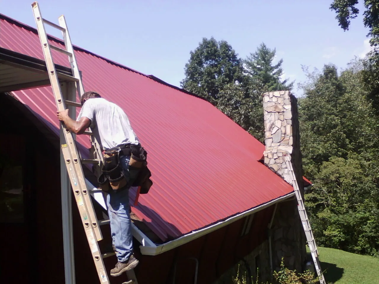 Roofer on a ladder beside a red metal roof and stone chimney — Soffit & Fascia Repair by Roofing Brothers in Meansville